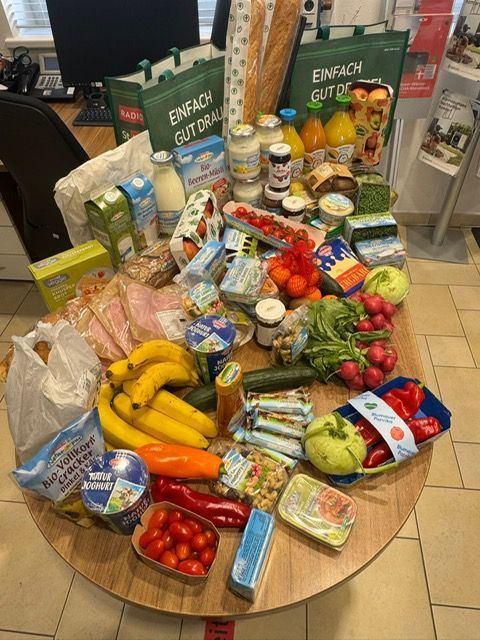 Auf einem runden Tisch ausgebreitet: Gemüse, Obst, Milchprodukte, Brot, Fleisch und wiederverwendbare Einkaufstaschen.