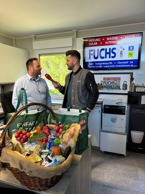 Eine Person interviewt eine andere in einem Geschäft; neben ihr stehen ein Korb und Tüten mit Lebensmitteln. Hinter ihnen ist ein Schild zu sehen.