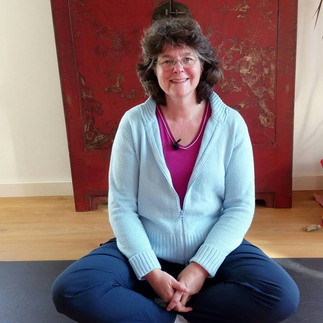 Een vrouw in een lichtblauw jasje zit met gekruiste benen glimlachend voor een rode kast.