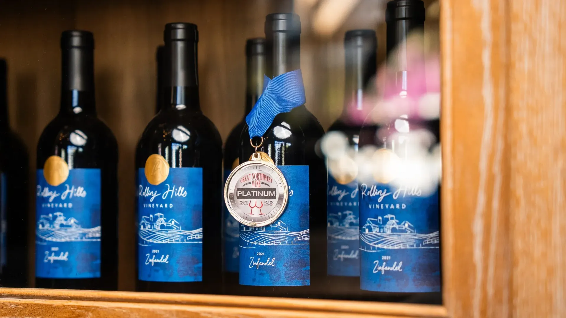 Bottles of wine on a shelf, one with a medal. Blue labels, gold accents, in a wooden cabinet.