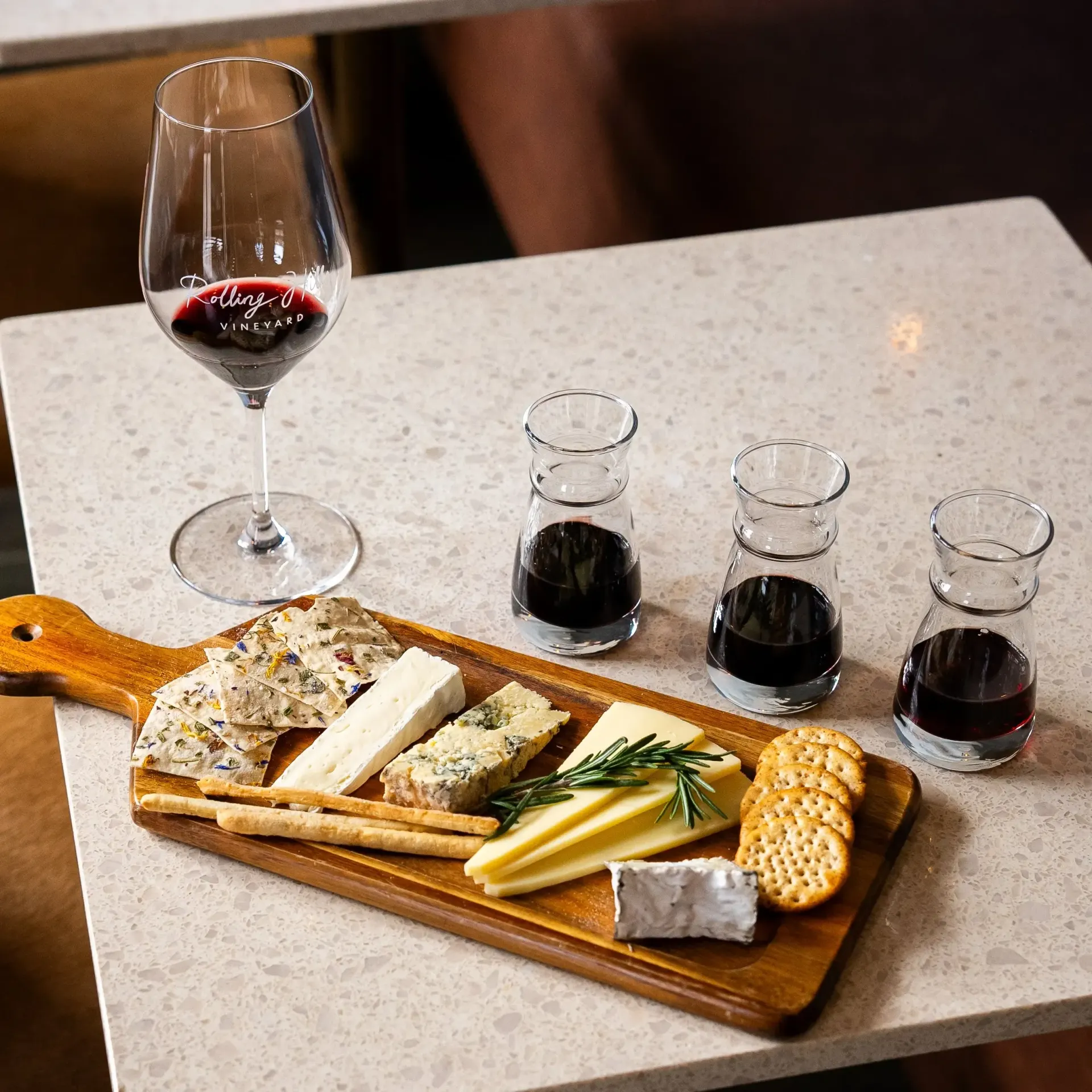 Wooden board with cheeses, crackers, and wine glasses on a light-colored table.
