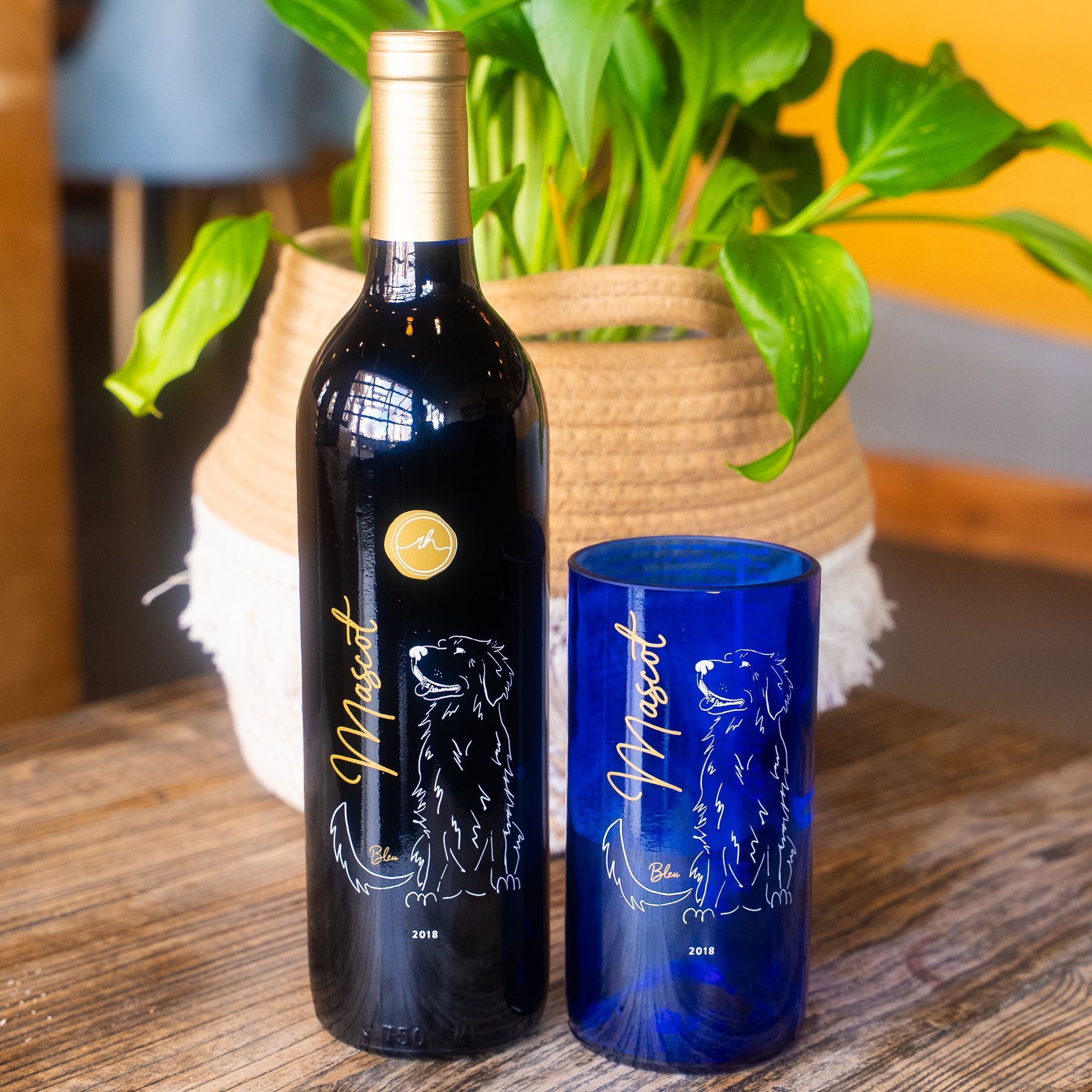 Wine bottle and glass with dog illustration, blue and gold, on a wooden table, with a plant in a basket.