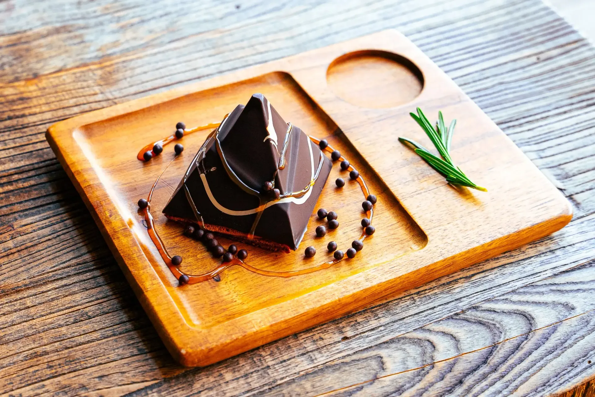 Chocolate pyramid dessert on wooden tray, drizzled syrup and peppercorns, sprig of herbs.