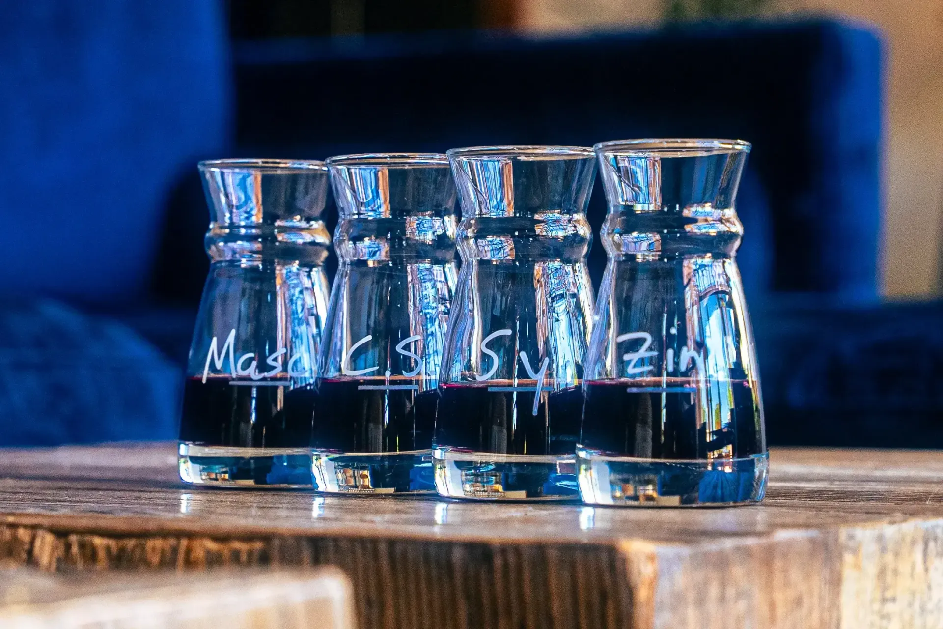 Six clear glass shot glasses with dark liquid, labeled on a wooden table.