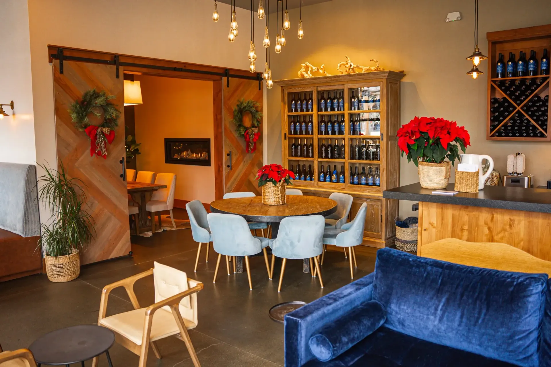 Cozy wine tasting room with round table, blue chairs, wooden cabinets, and Christmas decorations.
