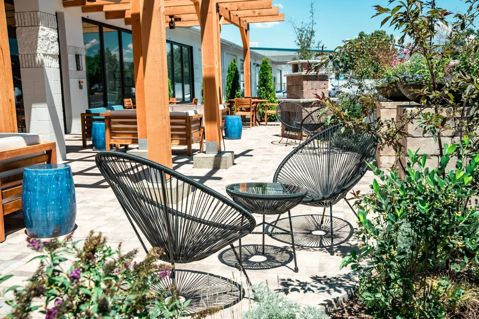 Outdoor patio with black woven chairs, small table, blue pots, wooden pergola, and greenery.