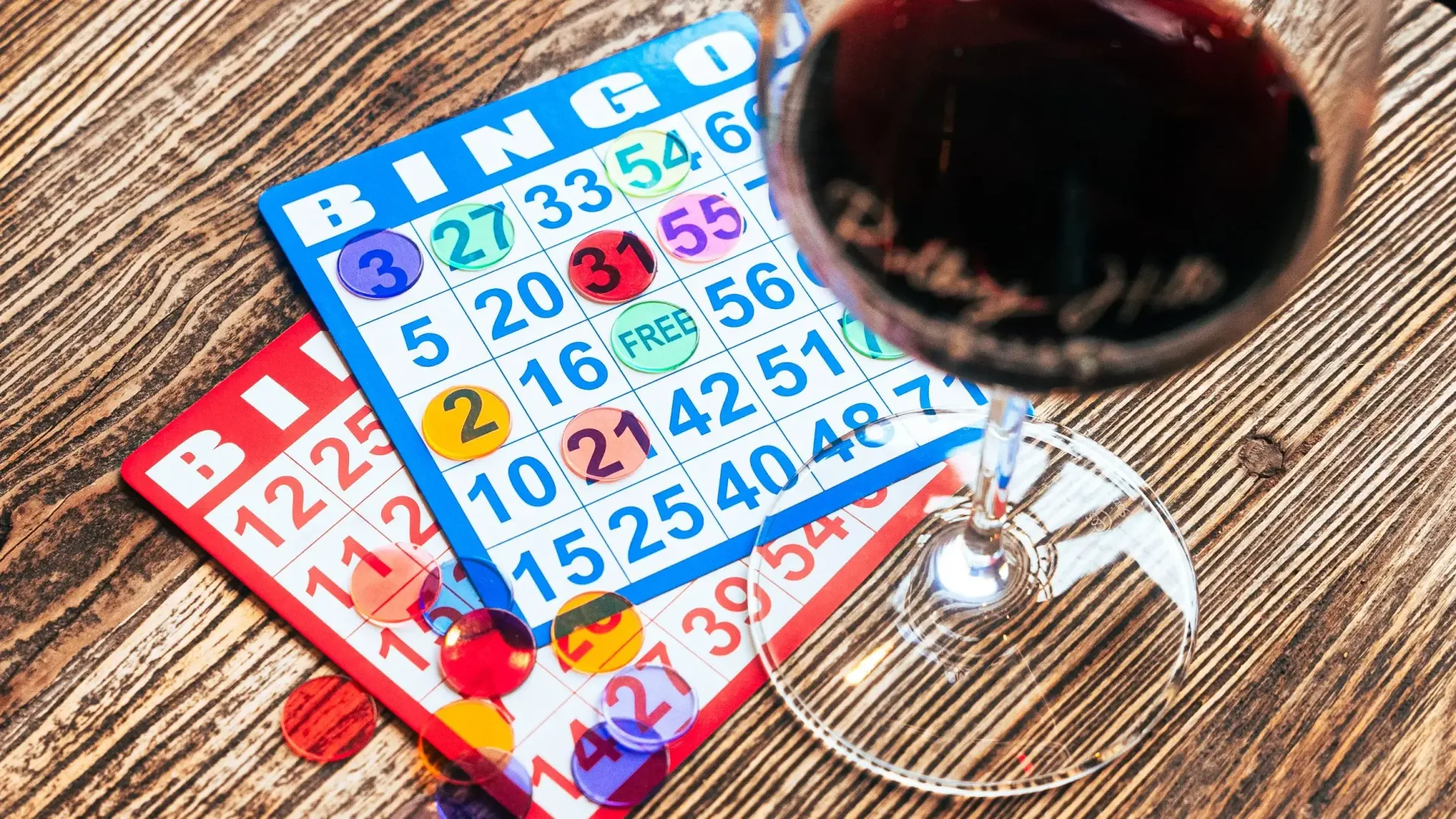 Two bingo cards with colorful markers and a glass of red wine on a wooden surface.