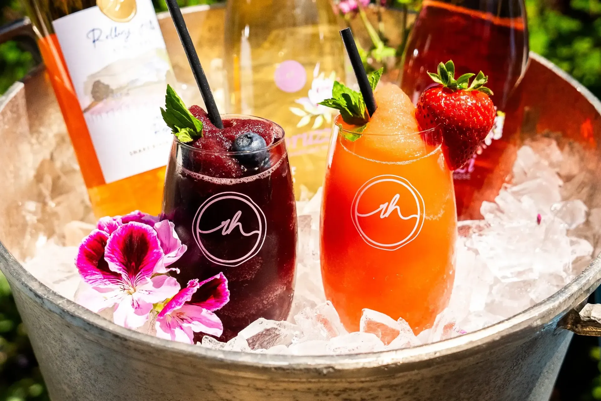 Cocktails in ice bucket, garnished with fruit and flowers, with bottles of wine in background.