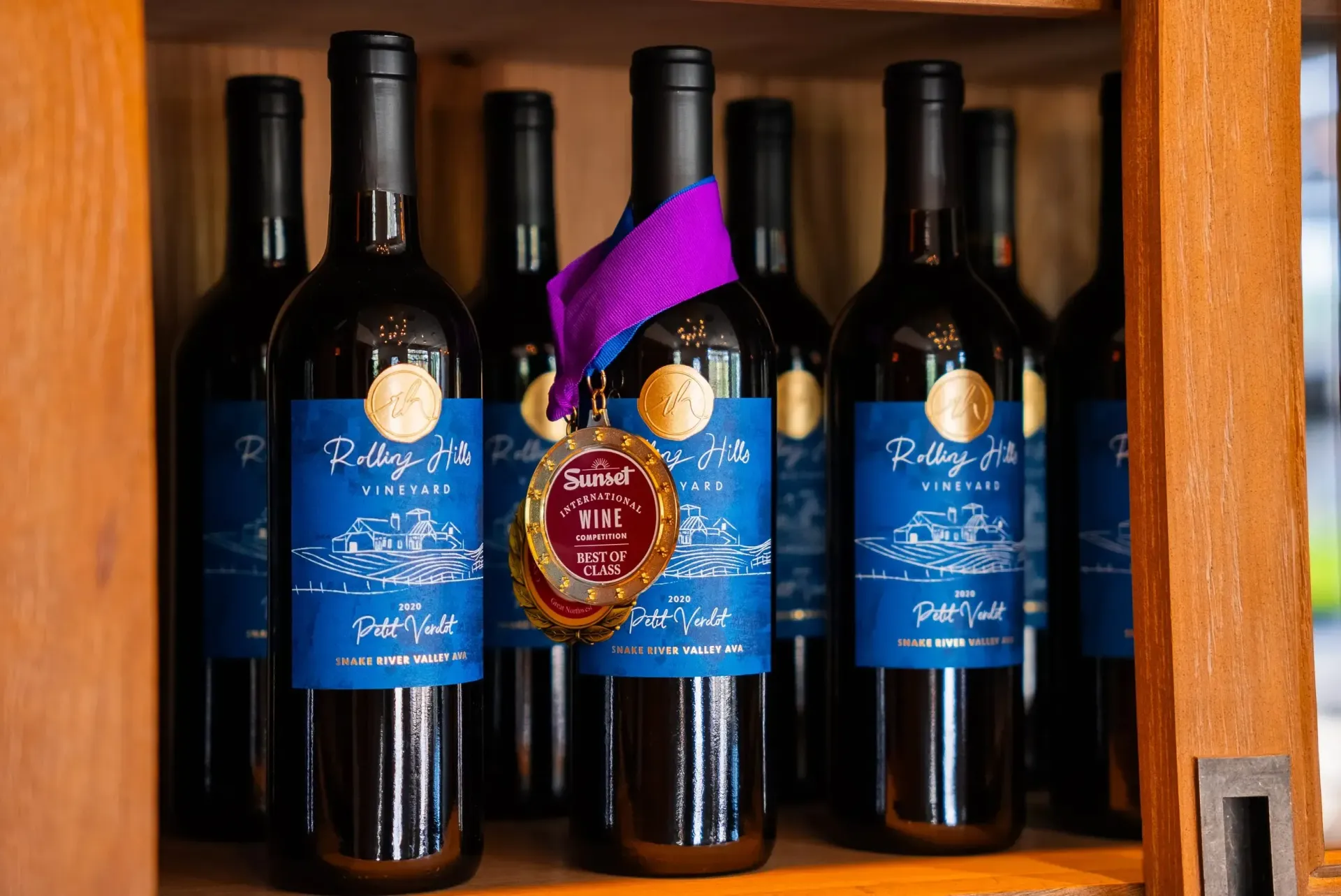 Bottles of red wine with dark blue labels and a gold medal on one. Wooden shelf.