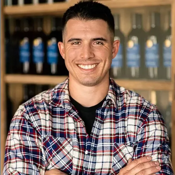 Man smiling with arms crossed, wearing a plaid shirt and standing in front of shelves of bottles.