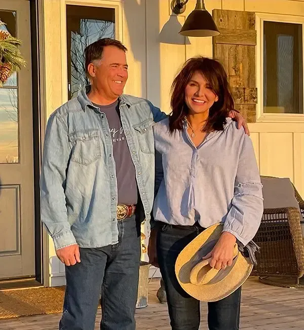 Couple outdoors, smiling, standing close. Man in denim shirt. Woman holds a hat, arm around man. Wooden wall backdrop.