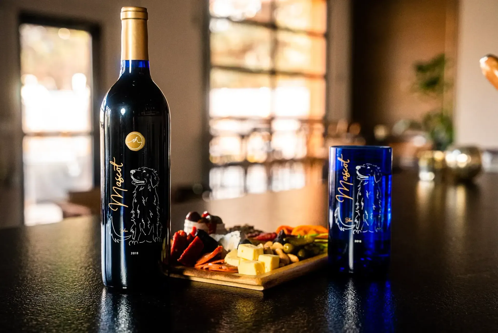 Dark blue wine bottle and glass with a charcuterie board on a black counter, inside.