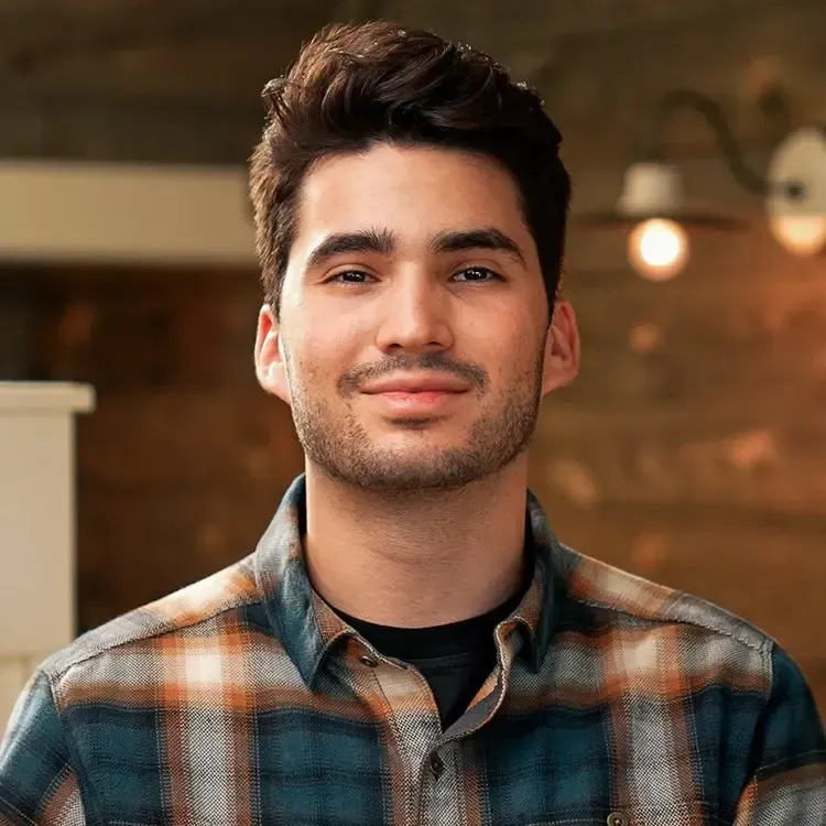 Man with dark hair and stubble, wearing a plaid shirt, smiling indoors.