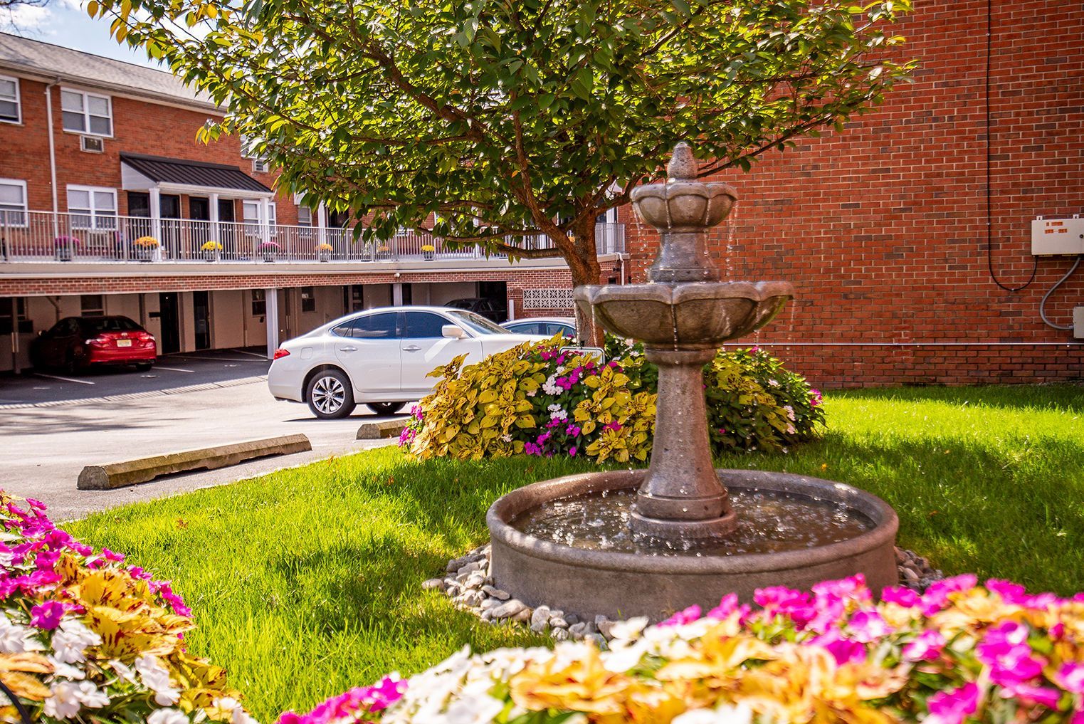 Exterior courtyard with a stone fountain, flowering shrubs, and parked cars.