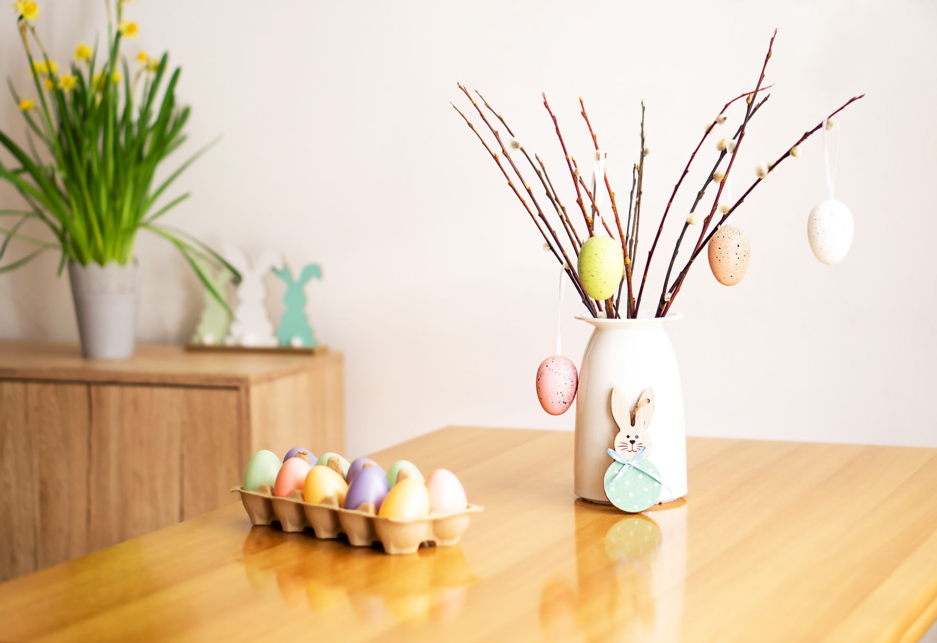 Easter decorations on a wooden table: eggs, a vase with branches and hanging eggs, and a potted plant.