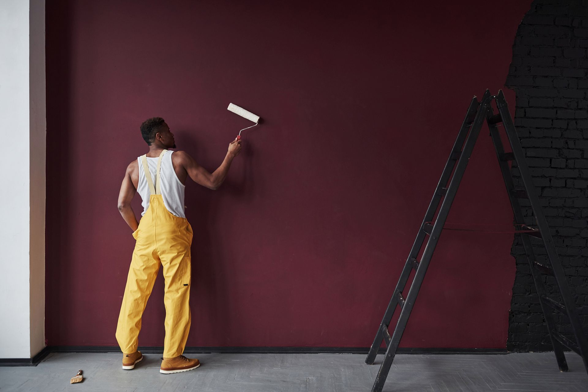 Man in yellow overalls paints a dark red wall with a roller, black ladder beside him.