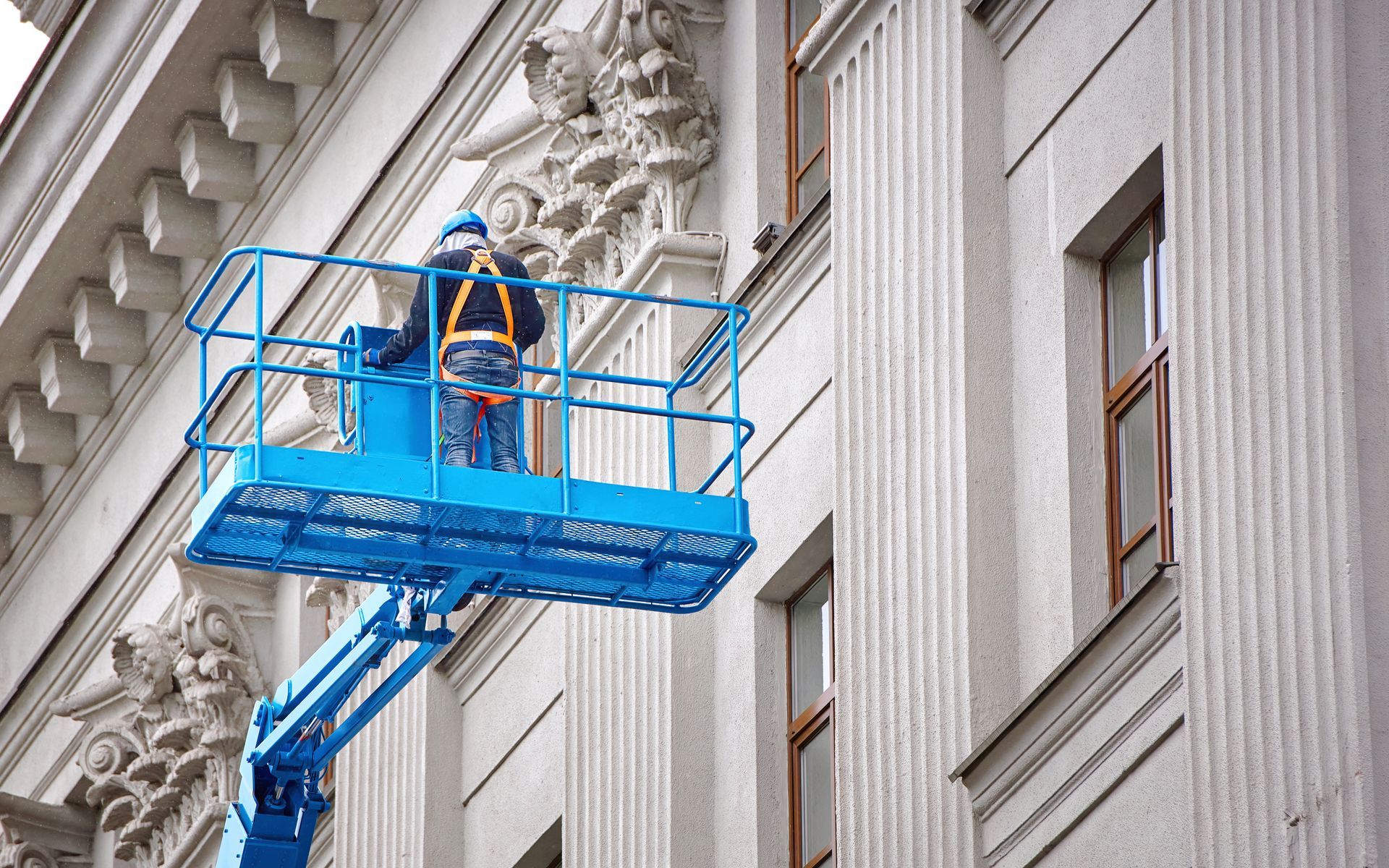 Worker in a blue lift inspecting ornate stonework on a building facade, wearing a safety harness.