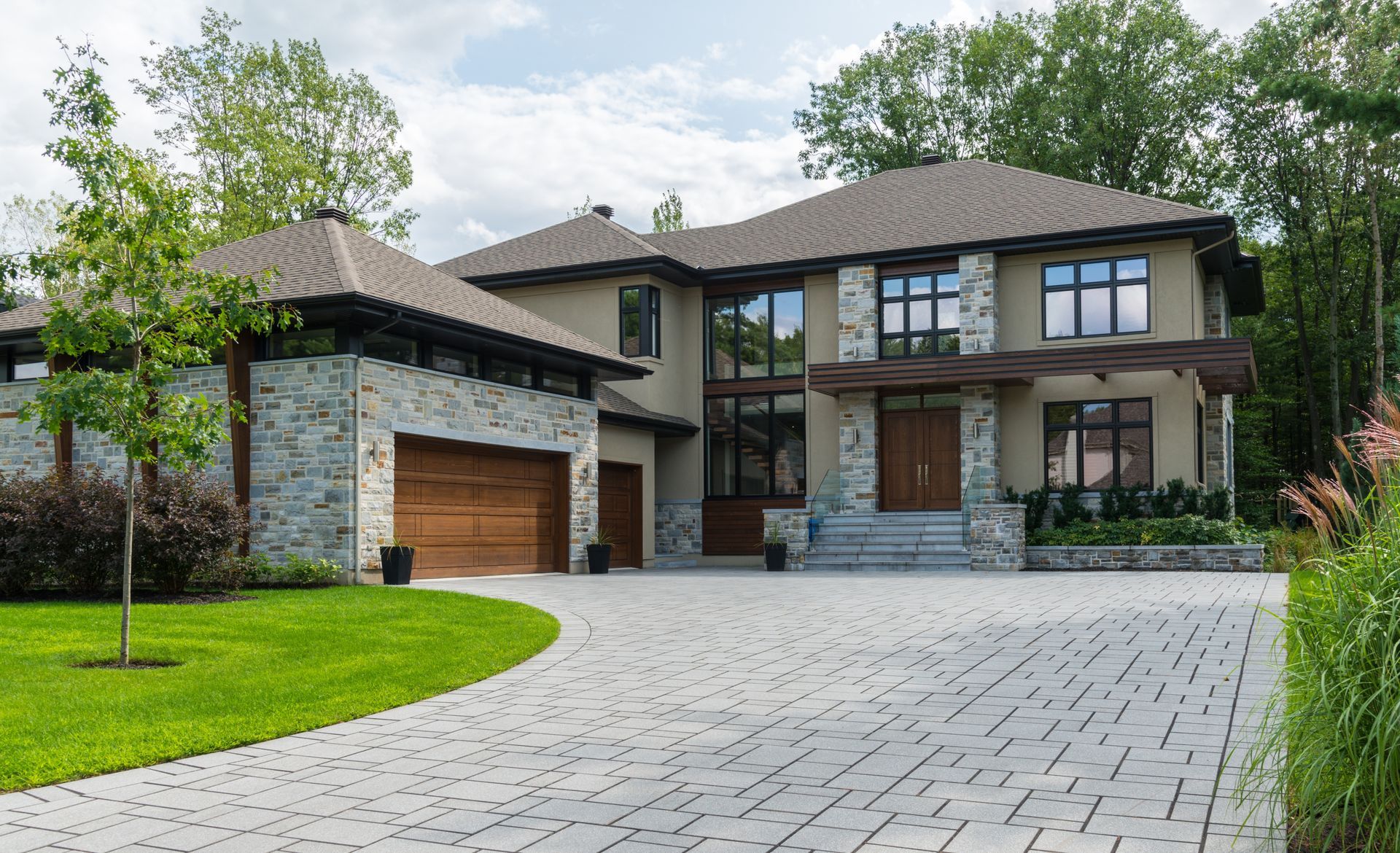 Modern house with stone and wood exterior, paved driveway, green lawn, and surrounding trees.
