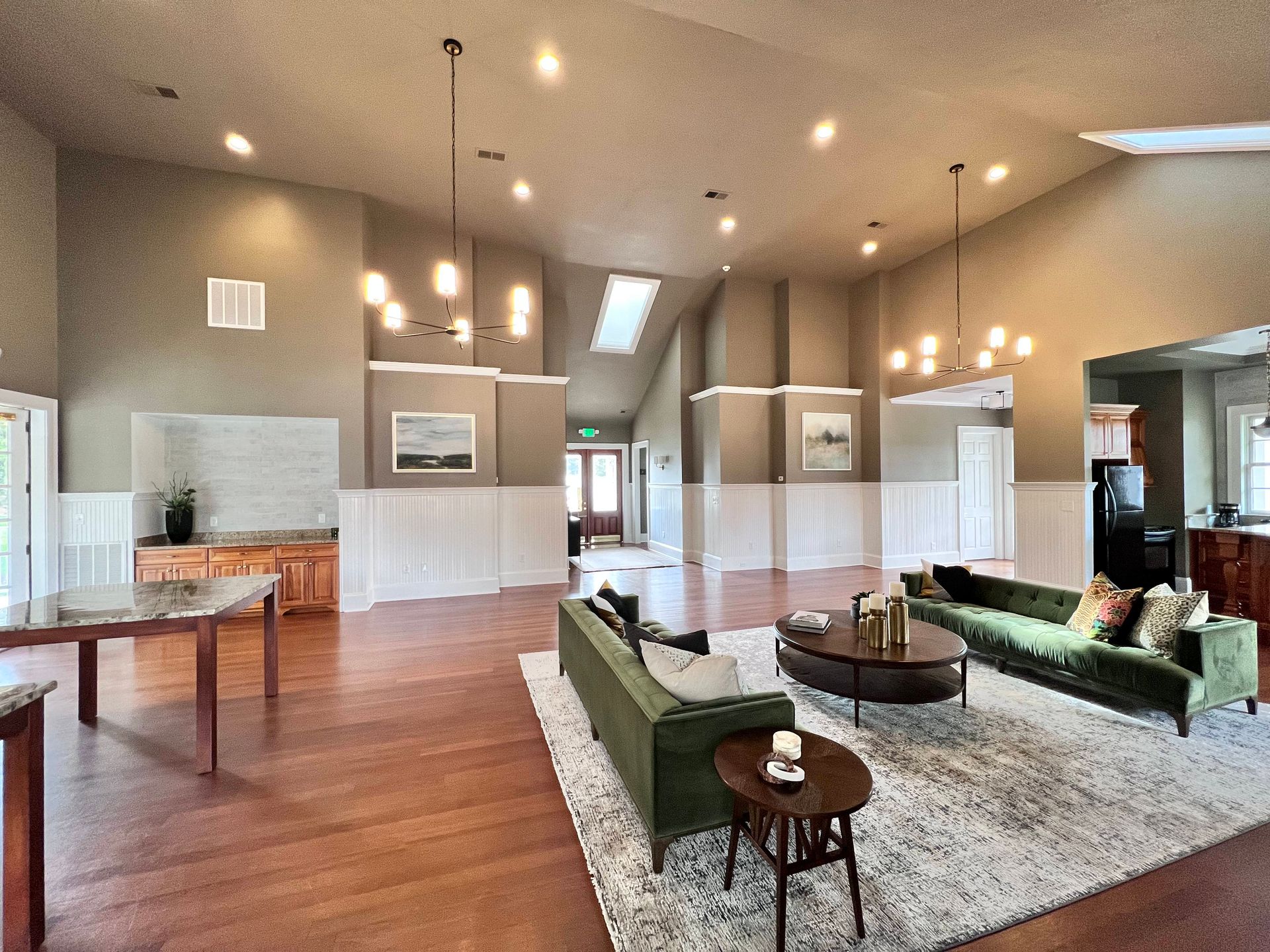 Spacious living room with green sofas, wooden floors, and chandeliers; light streaming through a skylight.