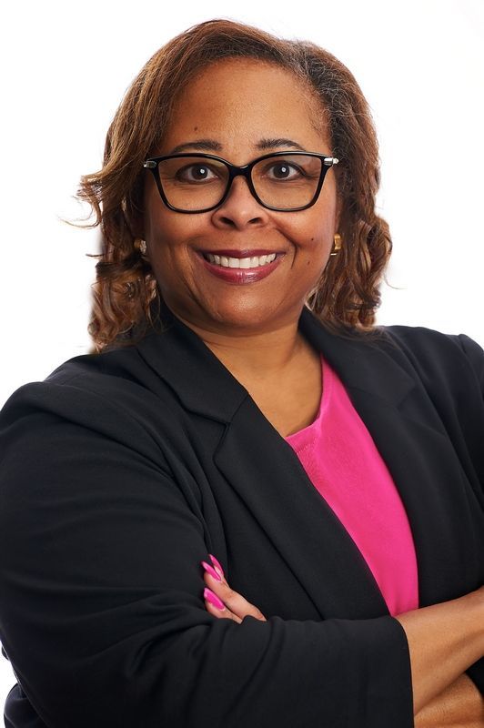 Deborah Lowery, co-founder of MINA wearing glasses and a black jacket is smiling with her arms crossed.