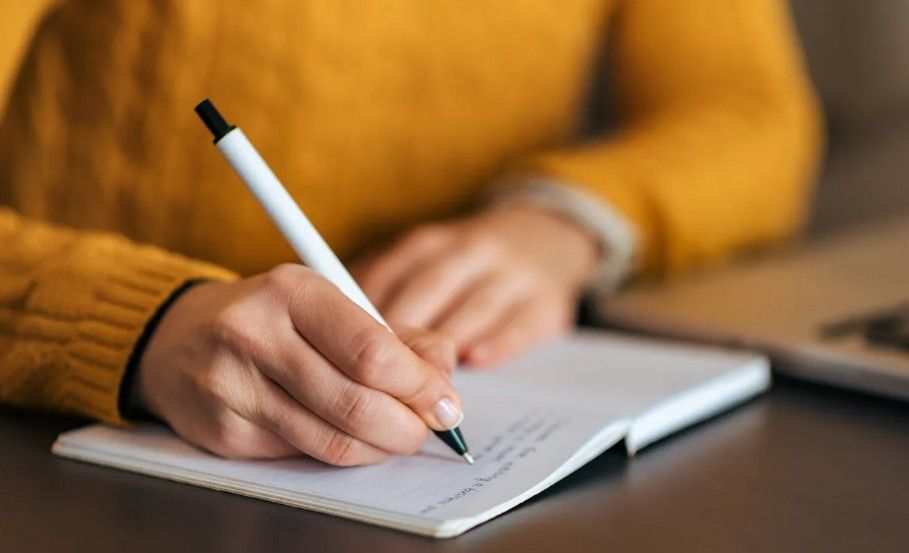 Woman working on laptop, taking notes at a desk in an office setting, smiling.