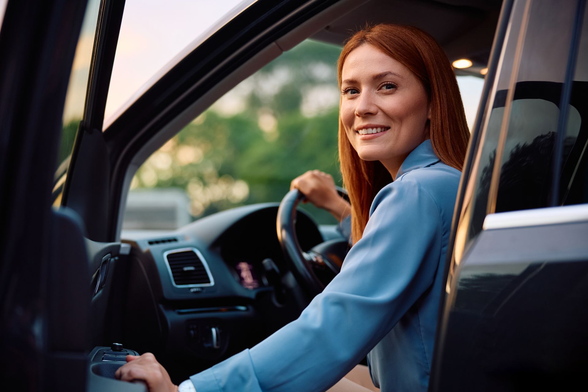 A smiling person sits in the driver's seat of a car, holding the steering wheel and looking back toward the camera.