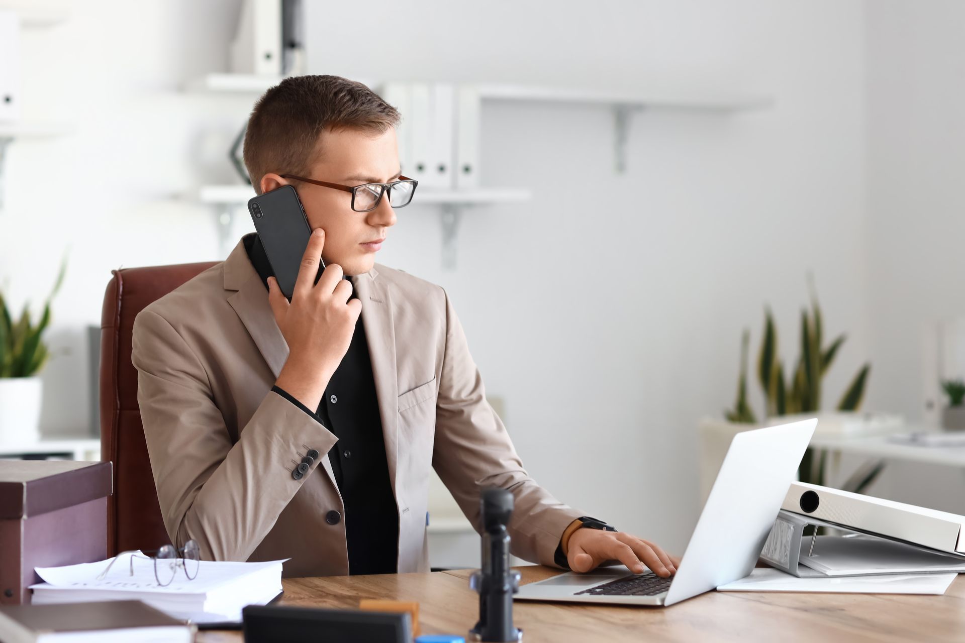 A man is sitting at a desk talking on a cell phone while using a laptop computer.