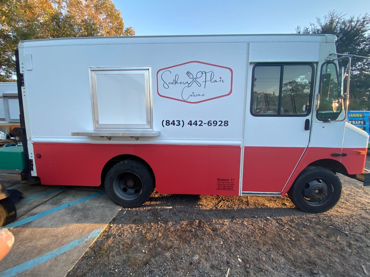 White and red food truck parked on gravel, with a service window and phone number on the side