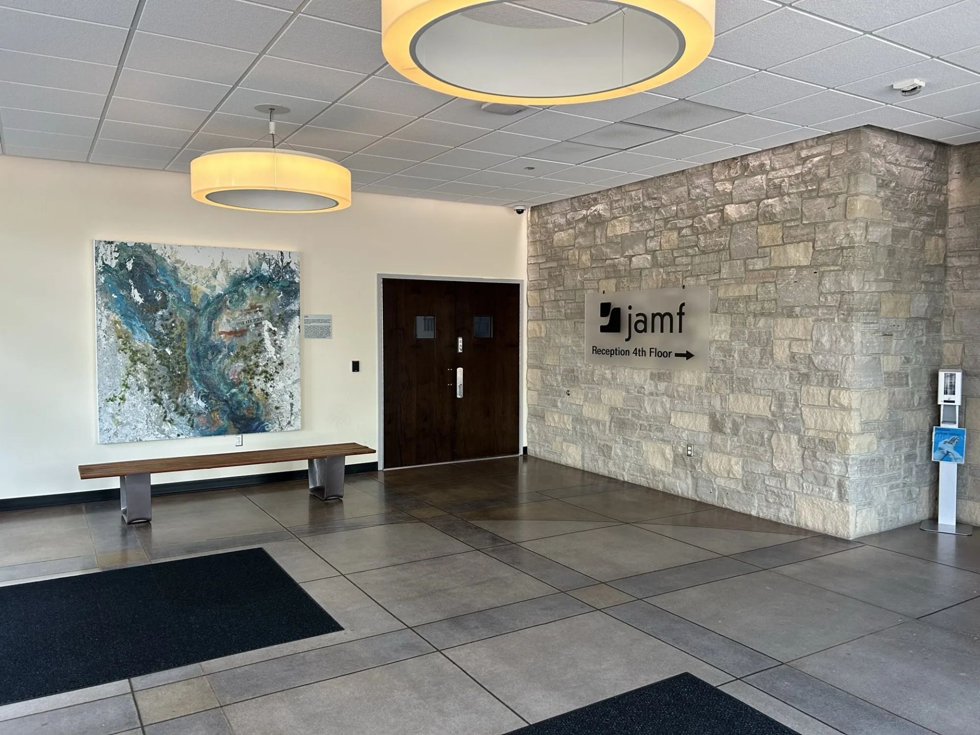 Lobby entrance with artwork, bench, and stone wall. Sign for jami, a direction indicator, and a hand sanitizer dispenser.