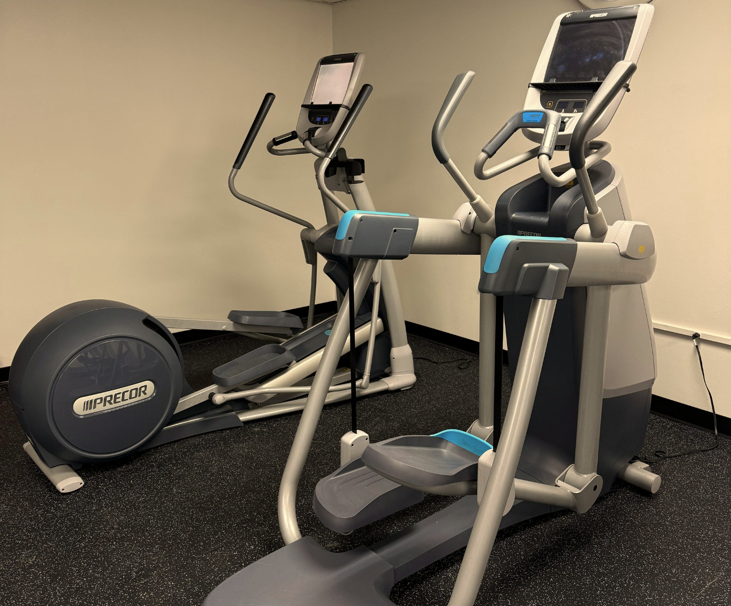 Two gray and blue elliptical machines in a gym setting on black flooring, against a tan wall.