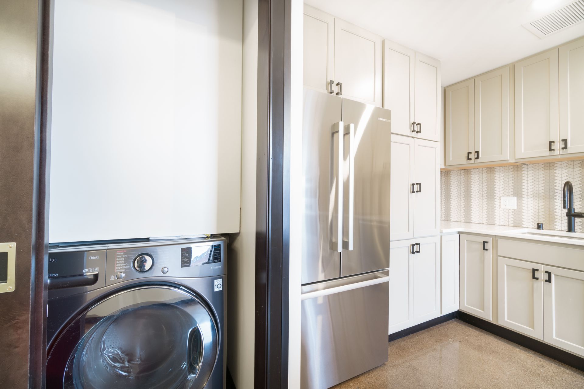 Washing machine in a closet, next to a kitchen with stainless steel refrigerator and white cabinets.