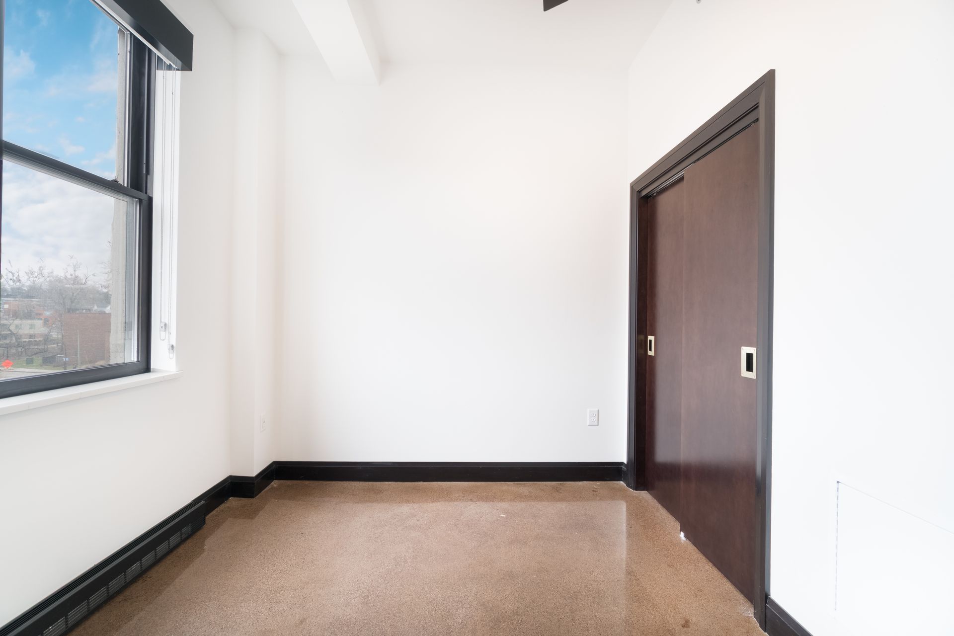 Empty room with window and closed brown door. White walls, dark baseboards, and light flooring.