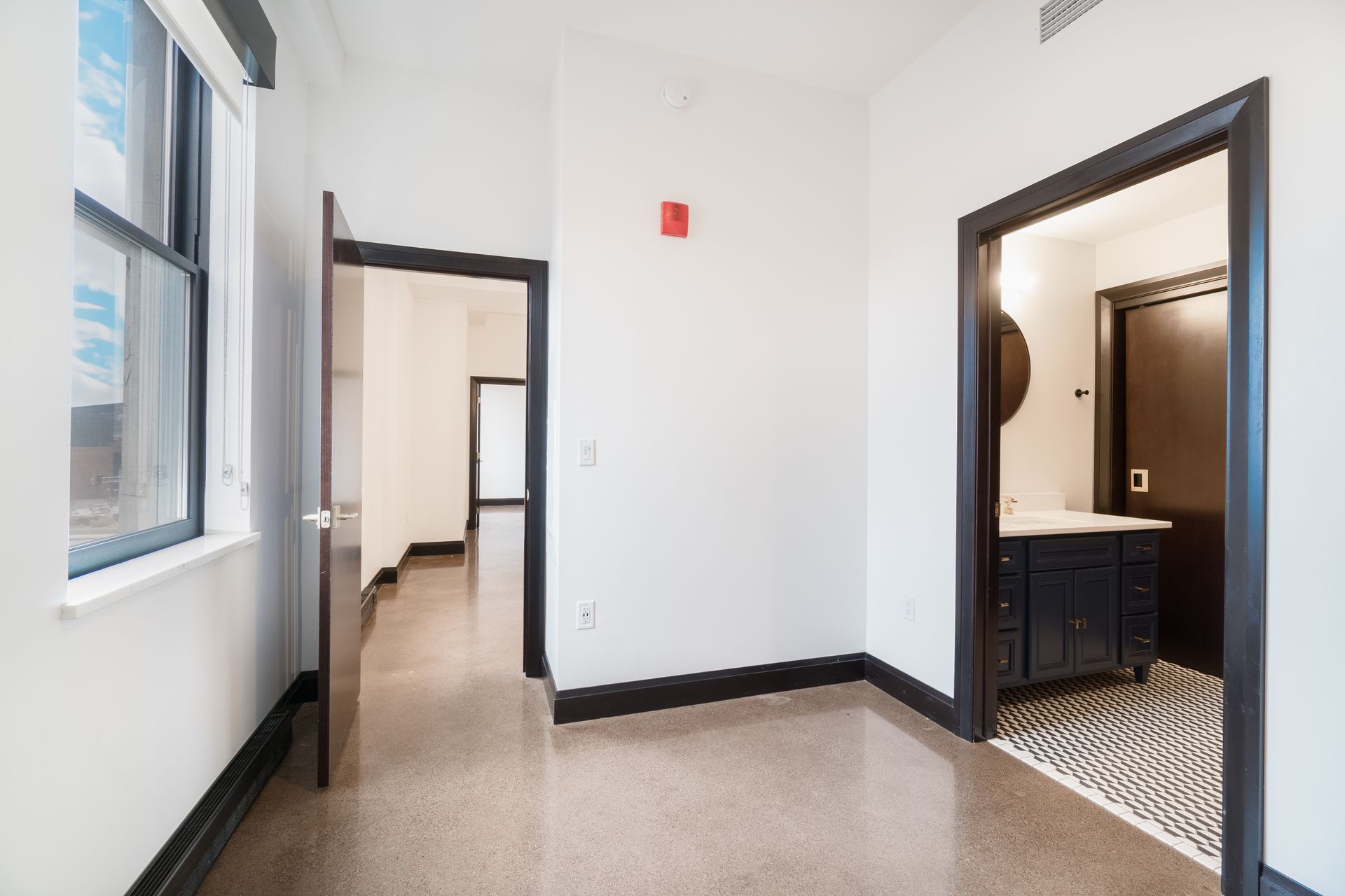 Empty room with doorways leading to bathroom, hall, and another room; window on the left; white walls, dark trim.