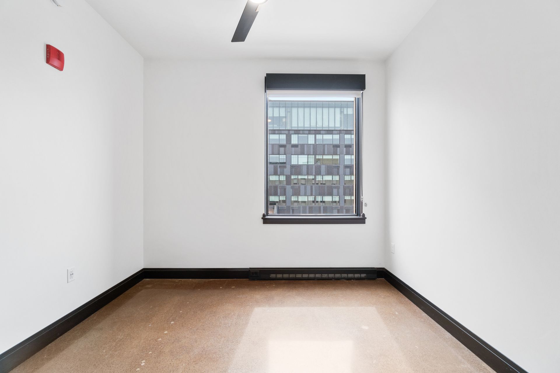Empty room with a window looking out onto a city building. White walls and brown floor.