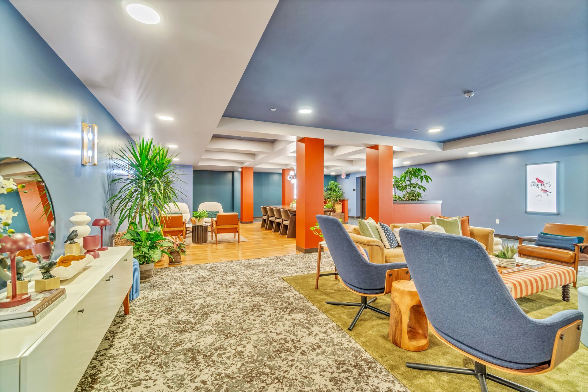 Lounge area with blue chairs, orange columns, and wooden tables. Light-colored rug and blue-gray walls.