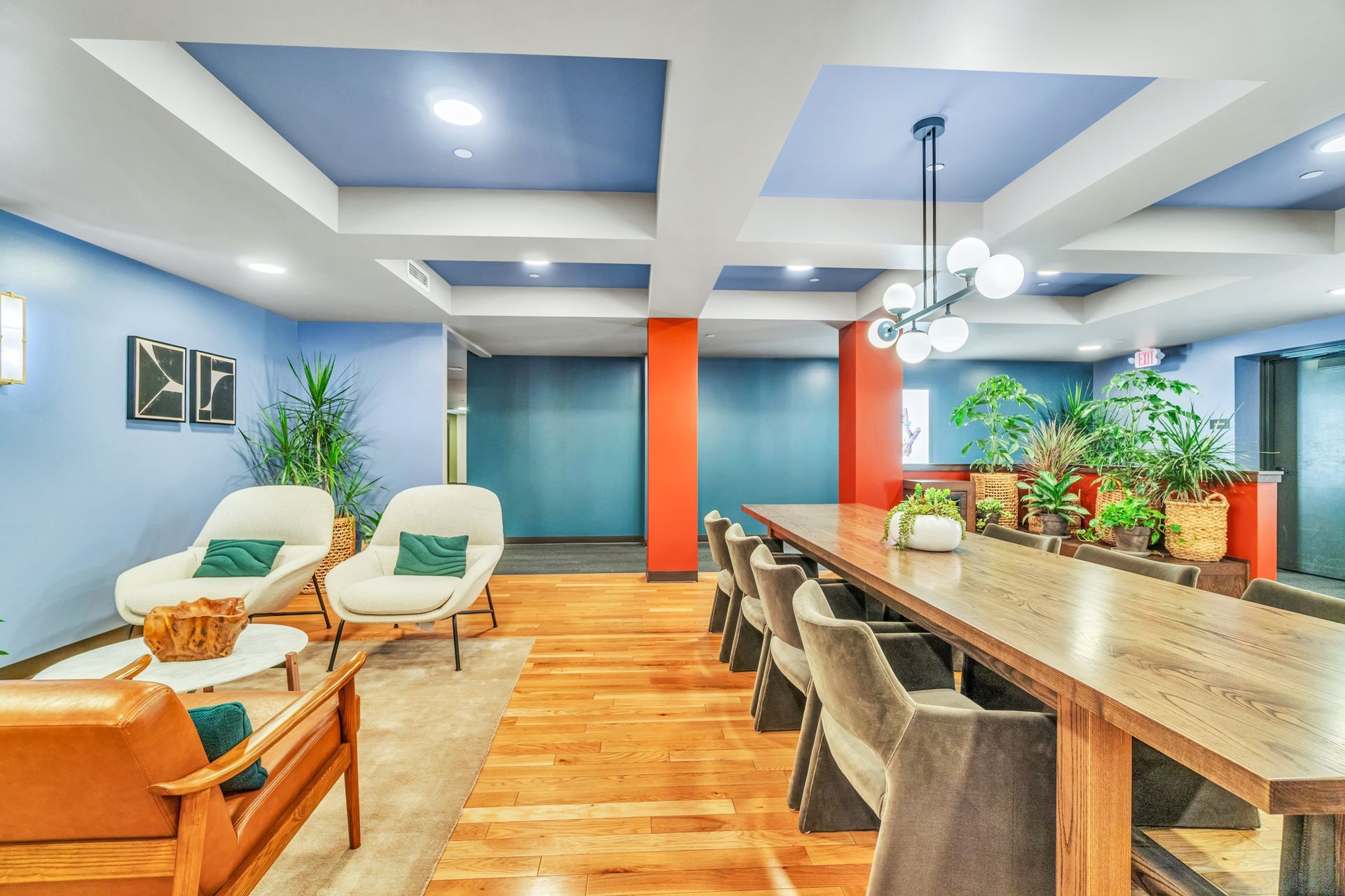 Bright lobby with seating: armchairs, long table, plants, wood floor, blue walls, and orange accents.