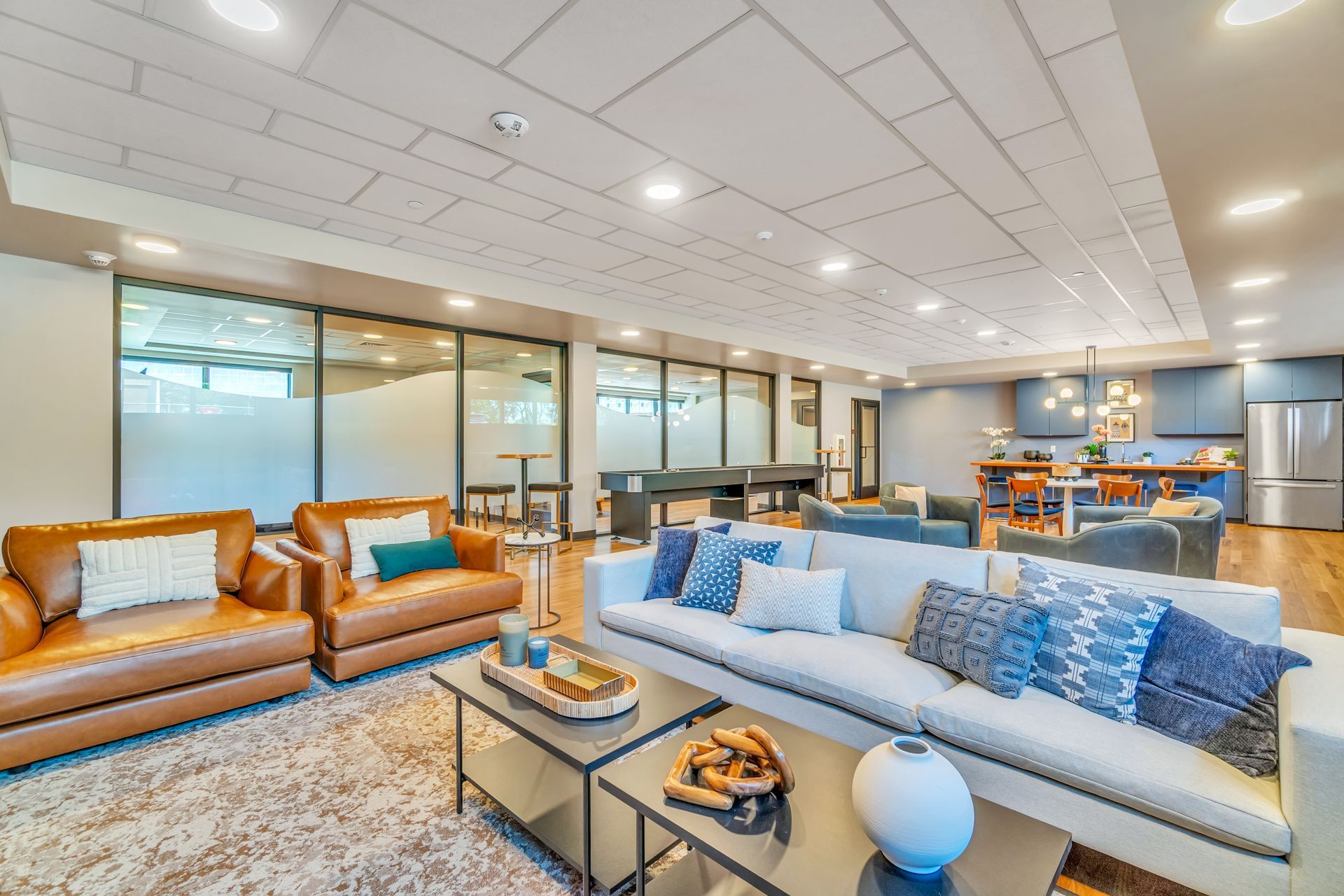 Spacious lounge with brown leather chairs, white sofa, coffee tables, and a kitchen in the background.