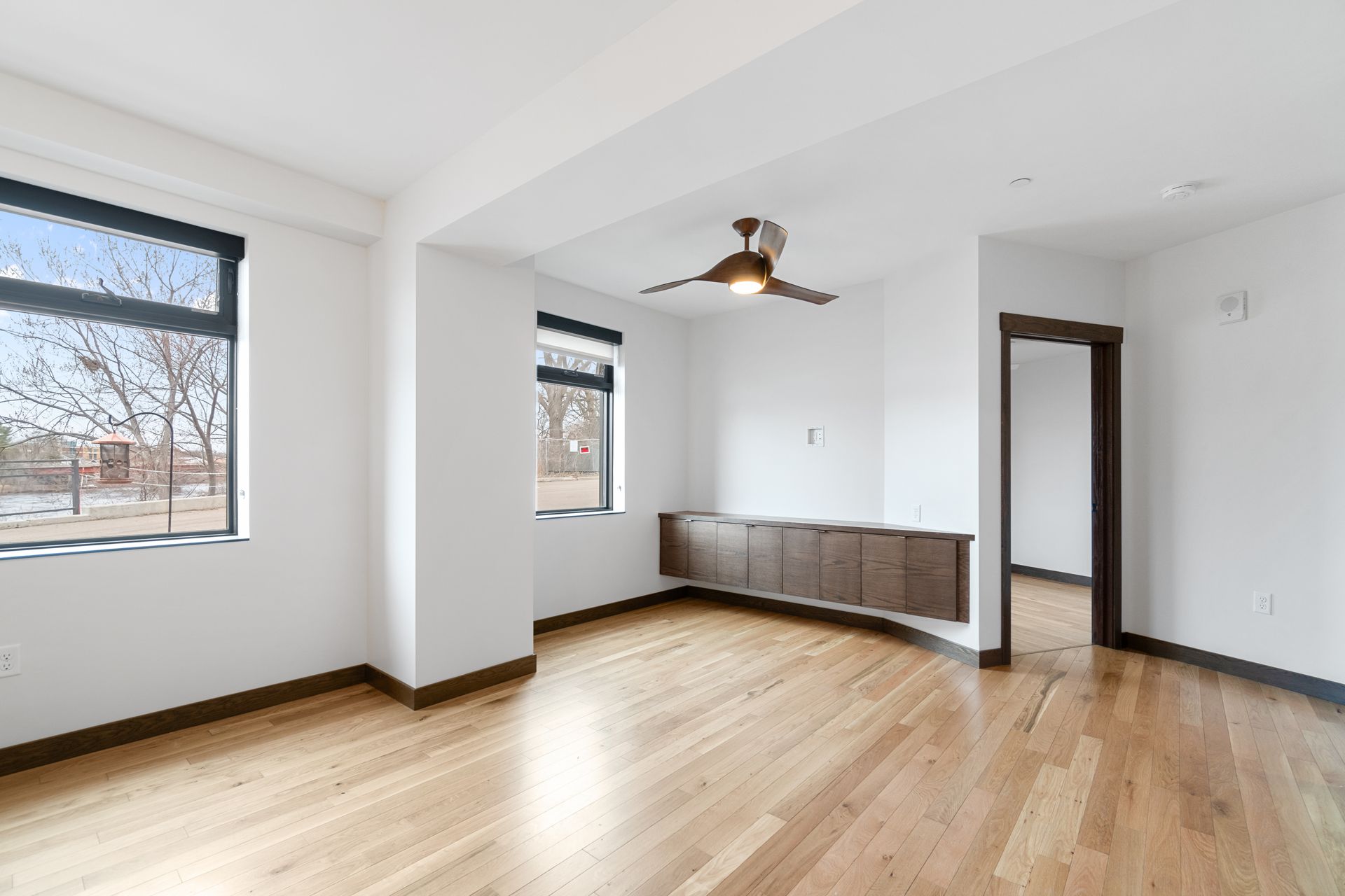 Bright, empty room with hardwood floors, white walls, and windows.  A built-in brown cabinet sits under a window.