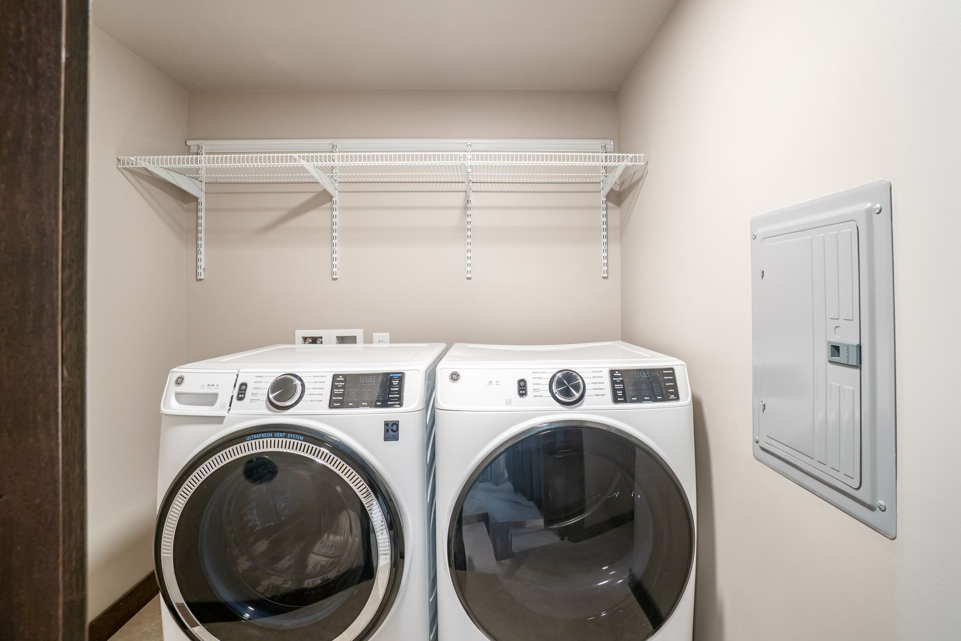 Laundry room with white washer and dryer, shelves above, and electrical panel on the wall.