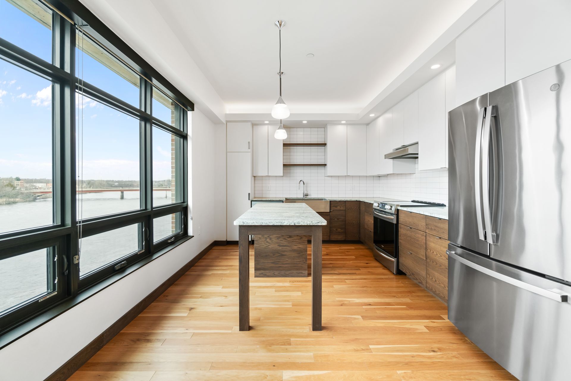Modern kitchen with large windows, hardwood floors, white cabinets, and a stainless steel refrigerator.