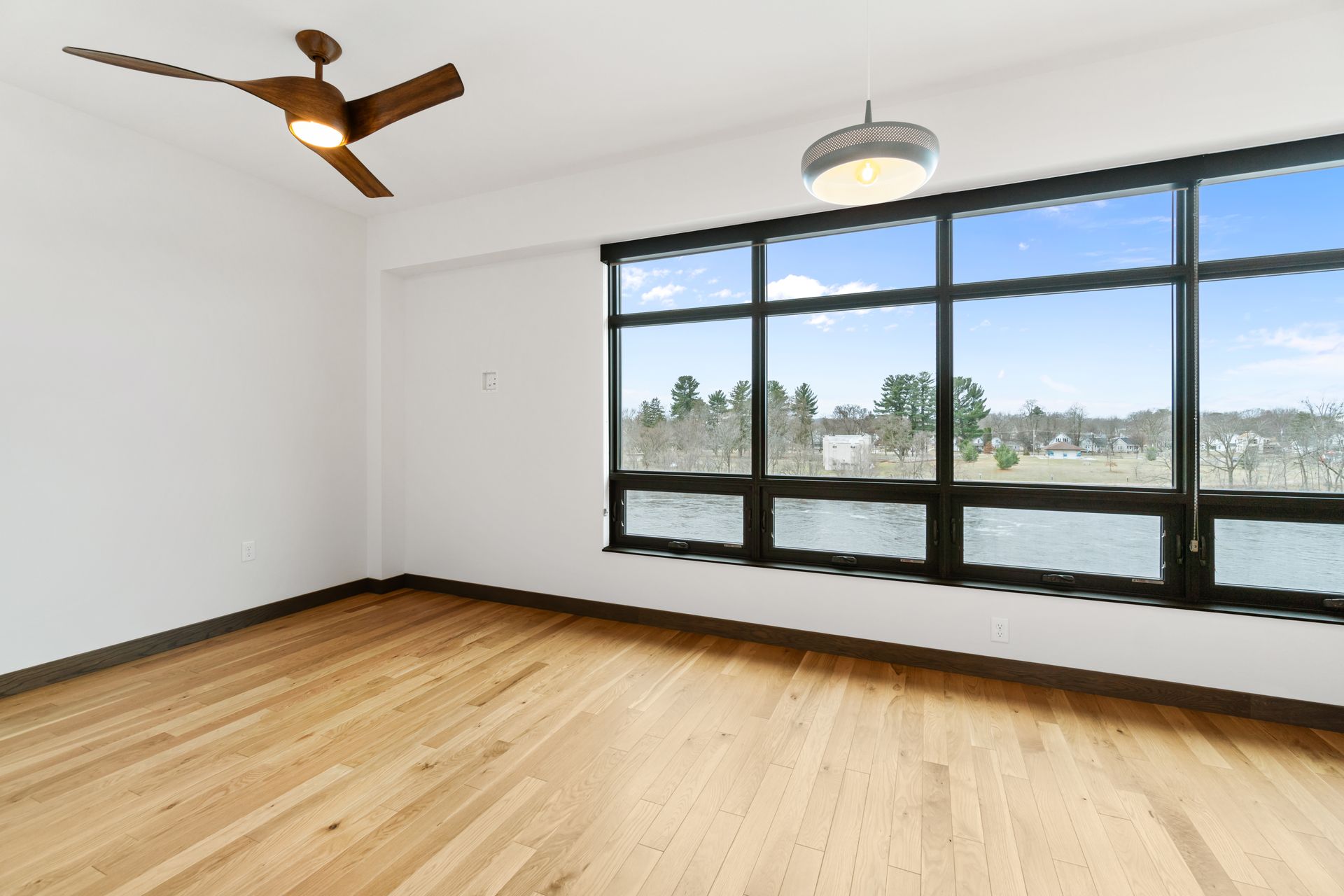 Empty room with wood floor, large window with outdoor view, ceiling fan and light fixture.