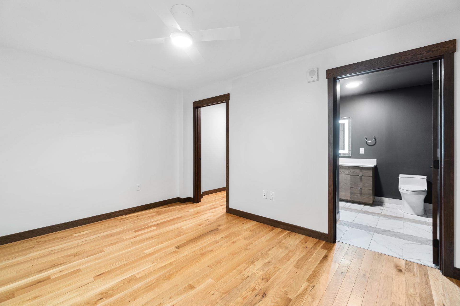 Empty bedroom with wood floors, two brown-framed doorways, and a bathroom visible.