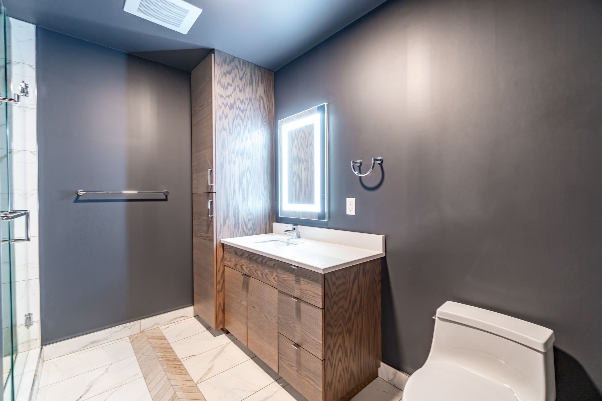 Bathroom with dark gray walls, wooden vanity, lighted mirror, and white toilet.