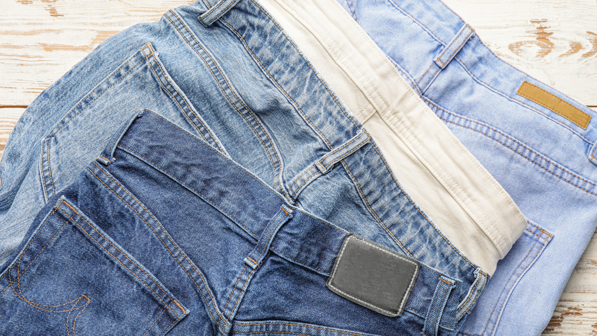 Three pairs of jeans are stacked on top of each other on a wooden table.