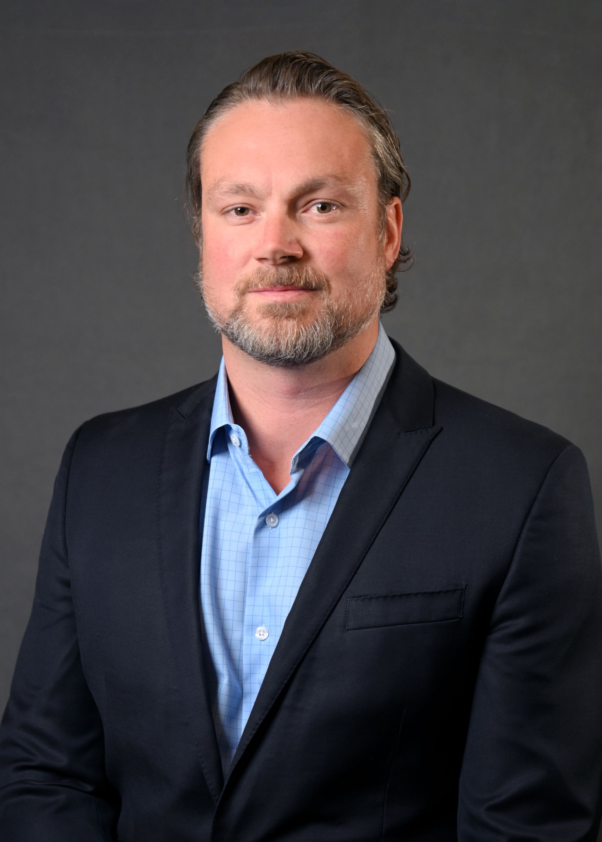 Man in a navy blazer and blue shirt smiles, looking at the viewer, against a grey background.
