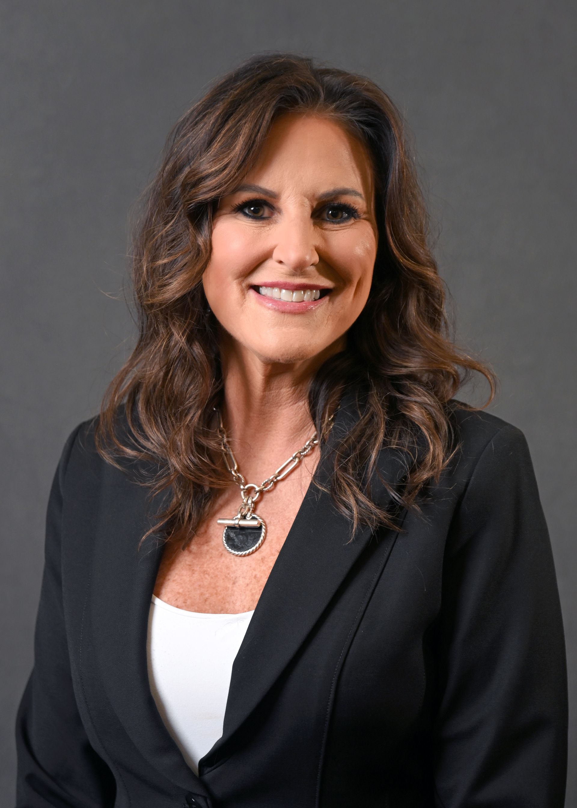 Woman with brown hair in a black blazer and white top, smiling, against a gray backdrop.