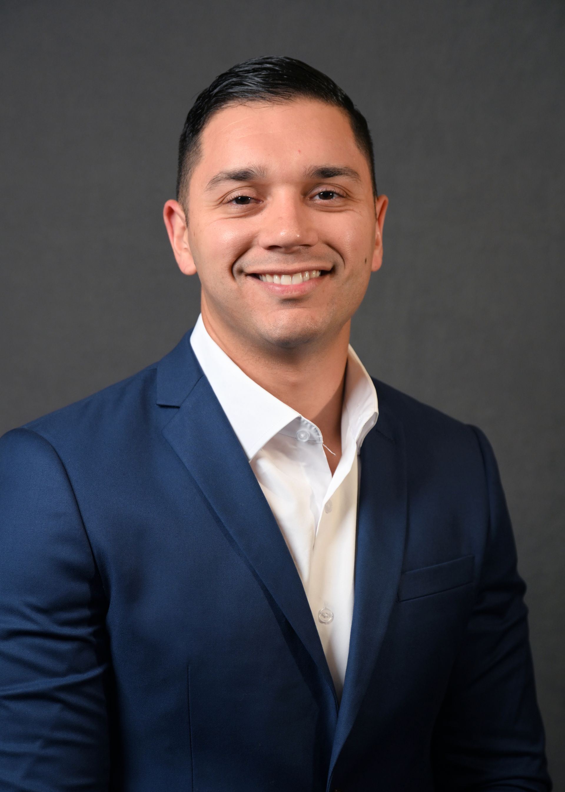 Man in a blue suit and white shirt smiling at the camera against a gray background.