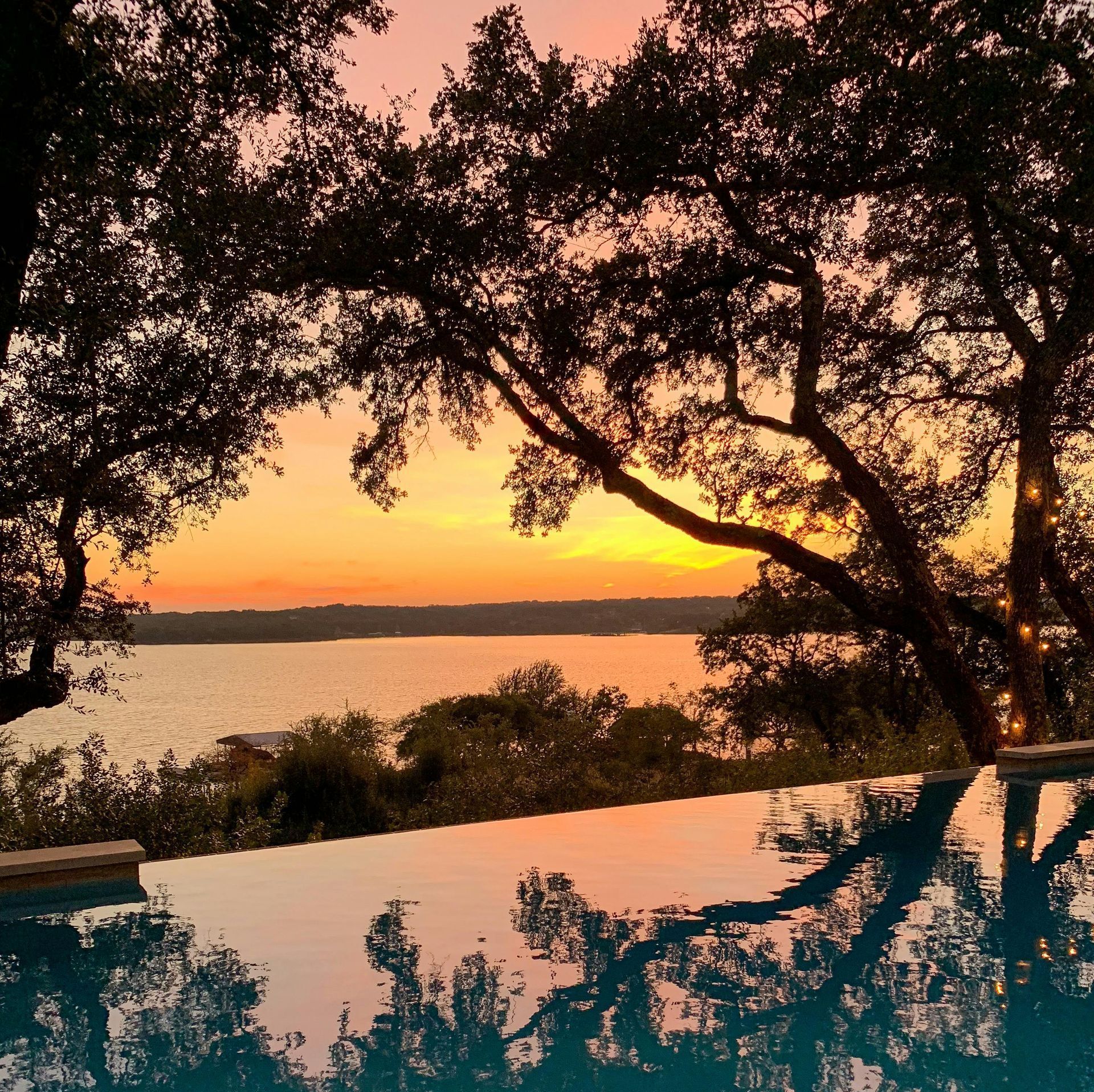 Sunset over a lake, reflected in an infinity pool, framed by silhouetted trees. Orange and pink sky.