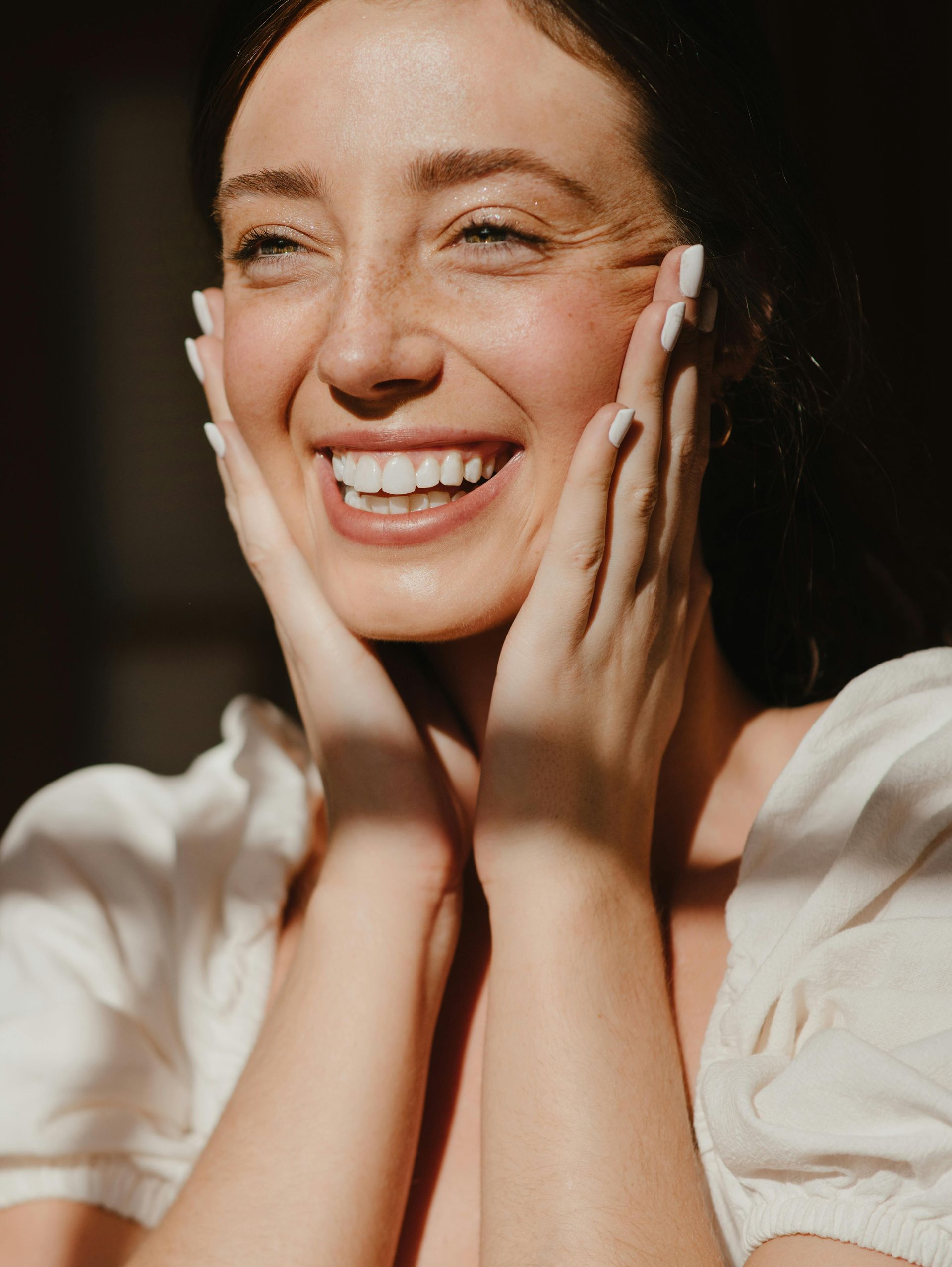 Woman smiling, hands on face; freckles; sunlight.