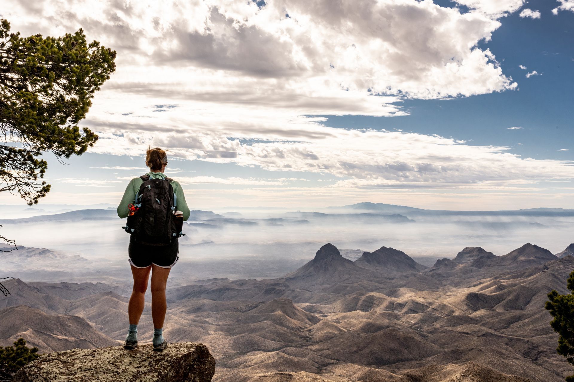 Woman with backpack stands on a cliff overlooking a mountain range under a cloudy sky.